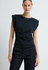 Top nero senza maniche con vita aderente, design drappeggiato e scollatura rotonda accentuata da un dettaglio in catena argentata. Abbinato a pantaloni in denim scuro.