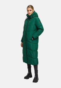 Woman wearing a long green padded winter coat with hood, black zipper, side pockets, black pants, and black chunky boots standing sideways.