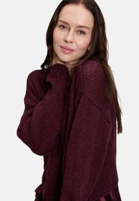Femme aux longs cheveux bruns portant un pull en laine ample et texturé de couleur violet foncé, tenant délicatement le col près de son visage contre un fond neutre.
