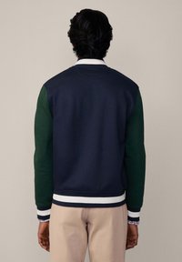 Marineblauwe sweatshirt met groene mouwen, ribgebreide manchetten en een witte gestreepte onderrand. Kenmerkt zich door een ronde halslijn en een logo-detail op de achterkant in groen.