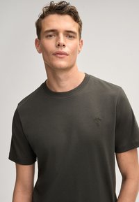 Dunkelgrünes Rundhals-T-Shirt aus weicher Baumwolle, mit kurzen Ärmeln und einem kleinen gestickten Design auf der Vorderseite.