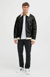 Jachetă din piele artificială neagră cu guler și manșete din blană crem, asociată cu pantaloni cargo negri și adidași albi. Design simplu și modern.
