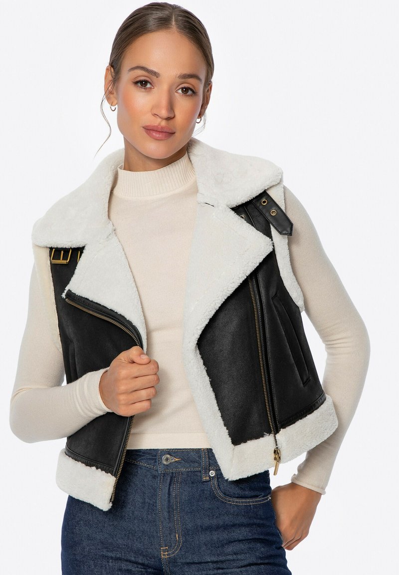Chaleco de cuero negro con cuello y forro de piel sintética blanca. Presenta detalles de cremallera y hebillas. El modelo lleva un suéter de cuello alto crema y pantalones vaqueros.