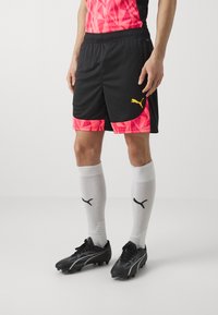 Pantaloni sportivi neri con un motivo geometrico rosa sull'orlo, abbinati a calzini bianchi con logo Puma neri e scarpini da calcio neri.