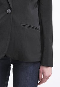 Blazer noir à bouton unique, coupe ajustée, avec une texture de tissu brossé. Comprend une poche latérale et des revers lisses.