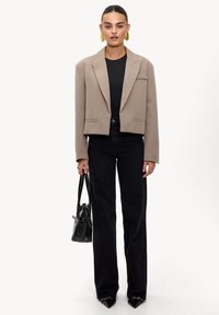 Blazer beige con rayas verticales cortado y cuello con muesca, combinado con jeans negros ajustados. La modelo lleva un bolso negro pequeño.