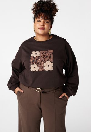 Femme aux cheveux bouclés portant des boucles d'oreilles en feuille d'or, un sweat-shirt à motifs floraux brun foncé et un pantalon marron assorti avec ceinture, les mains dans les poches.