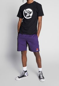 Musta T-paita, jossa on pyöreä lintukuva, yhdistettynä purppuranvärisiin shortseihin, joissa on punainen logopläjäys vasemmassa lahkeessa, ja mustat korkeavartiset tennarit.