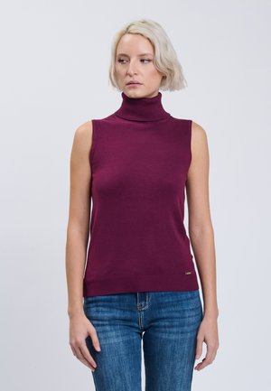 Frau mit kurzem blondem Haar trägt einen ärmellosen lila Rollkragenpullover und blaue Jeans und steht vor einem schlichten, hellen Hintergrund.