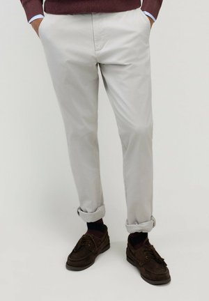 Hombre con pantalones beige claro remangados en los puños, zapatos marrón oscuro y un suéter granate con las manos en los bolsillos.