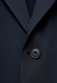 Zbliżenie na materiał marynarki w kolorze granatowym, z wyraźnym kołnierzykiem, gładką teksturą oraz okrągłym, czarnym guzikiem z czterema otworami o subtelnym połysku.
