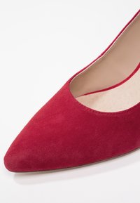 Chaussure à talon haut en daim rouge avec un bout pointu et des côtés découpés. Texture lisse et design minimaliste, mettant en valeur la teinte profonde.