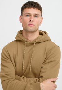 Khaki hooded sweatshirt with a drawstring and ribbed cuffs. Soft texture, relaxed fit, no visible patterns or logos. Model posing with arms crossed.