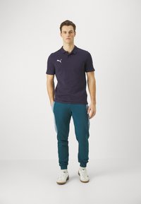 Marineblaues Poloshirt mit Kragen und weißem Logo, kombiniert mit türkisfarbenen Jogginghosen mit seitlichen Akzenten und weißen Sneakers. Das Model steht aufrecht.