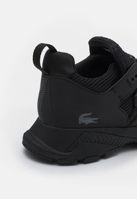 Black athletic shoe featuring a textured mesh upper, sleek rubber sole, and a grey crocodile logo on the side. Subtle stitching accents.