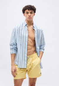 Νεαρός με σγουρά μαλλιά, φοράει ανοικτό μπλε ριγωτό πουκάμισο και κίτρινα μαγιό, με το χέρι στην τσέπη, μπροστά σε απλό φόντο.