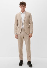 Lichtbeige kostuum met een aansluitend ontwerp, een een-knopsjas, bijpassende broek en een wit shirt. Zwarte sneakers maken de outfit compleet.
