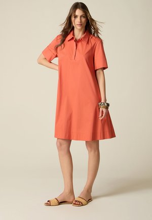 Donna che indossa un abito polo corallo fino al ginocchio con maniche corte, in piedi a piedi nudi in sandali intrecciati, accessoriata con bracciali spessi.