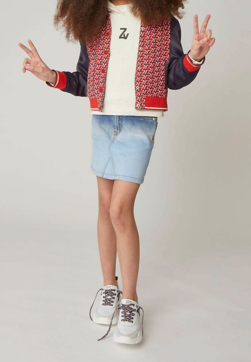 Blouson aviateur rouge et marine avec imprimé géométrique, sweat à capuche blanc avec logo, jupe en jean clair et baskets blanches avec lacets foncés.