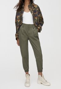 Veste à motif camouflage, pantalon vert olive avec un détail anneau, et bottes montantes beige. Tissu lisse, coupe décontractée, et poignets élastiques.