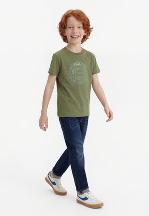 Usmievavý ryšavý chlapec kráča, má na sebe zelené tričko Moschino, tmavomodré džínsy a biele tenisky s modrými pruhmi na bielom pozadí.