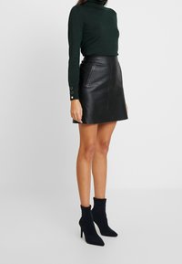 Schwarzer Leder-Minirock mit hohem Bund, kombiniert mit einem dunkelgrünen Rollkragenpullover und schwarzen Ankle Boots. Sichtbare strukturierte Stoffdetails.