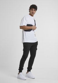 White oversized t-shirt paired with black cargo sweatpants and white sneakers. Model holds a black crossbody bag, standing against a simple background.
