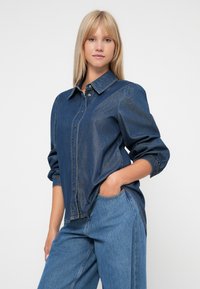 Denimskjorte i mørkeblått med knappeåpning, krage og puffermer. Kombinert med høyt liv denimbukser. Uformell passform.