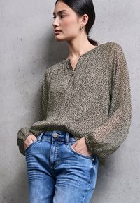 Letertil, langærmet bluse med grøn- og sort leopardmønster, parret med blå denimjeans, hænderne i lommerne, foran en betonvæg.