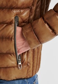 Braune Lederjacke mit gestepptem Muster, ausgestattet mit einer silbernen Reißverschlusstasche und einem weichen Bund, der ein figurbetontes Design und eine glatte Oberfläche hervorhebt.