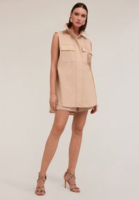 Abito beige senza maniche con colletto a punta e due tasche sul petto, abbinato a pantaloni crema e sandali con tacco a strappo. Tessuto dalla consistenza liscia.