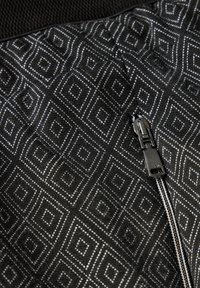 Un tessuto nero presenta un motivo geometrico a diamante grigio. Una cerniera con un lembo metallico è visibile, aggiungendo dettaglio al design.