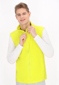 Neon žlutá bezrukávová vesta s vysokým límcem a p�ředním zipem, nošená přes přiléhavé bílé tričko s dlouhým rukávem. Hladká textura, neformální design.