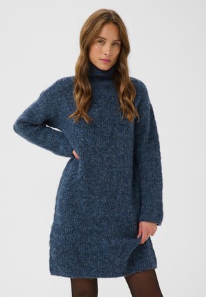 Femme aux longs cheveux ondulés porte une robe-pull à col roulé tricotée en poids bleu duveteux et des collants noirs, debout avec une main sur la hanche.