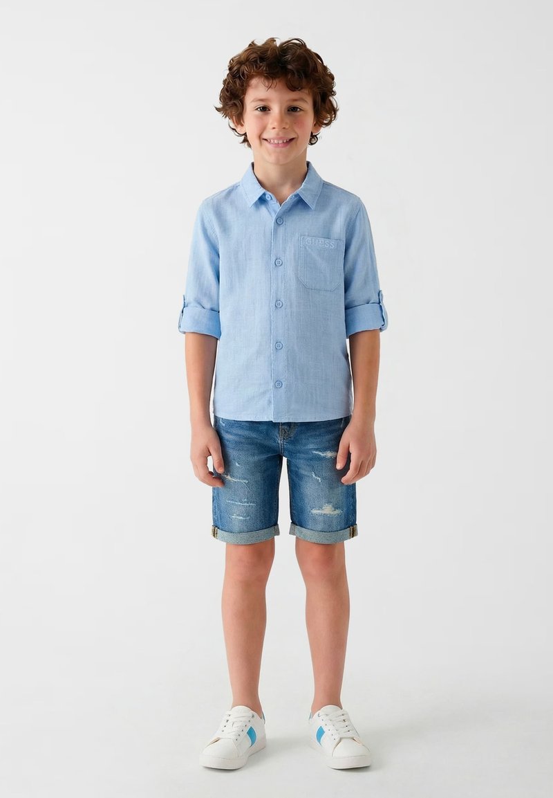 Garçon debout, portant une chemise bleu clair à manches longues boutonnée, manches retroussées, short en jean et baskets blanches avec des rayures bleues.