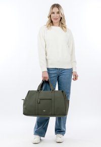 Vrouw draagt een witte trui, blauwe spijkerbroek en witte sneakers, en houdt een olijfgroene reistas met een voorvak met ritssluiting vast.