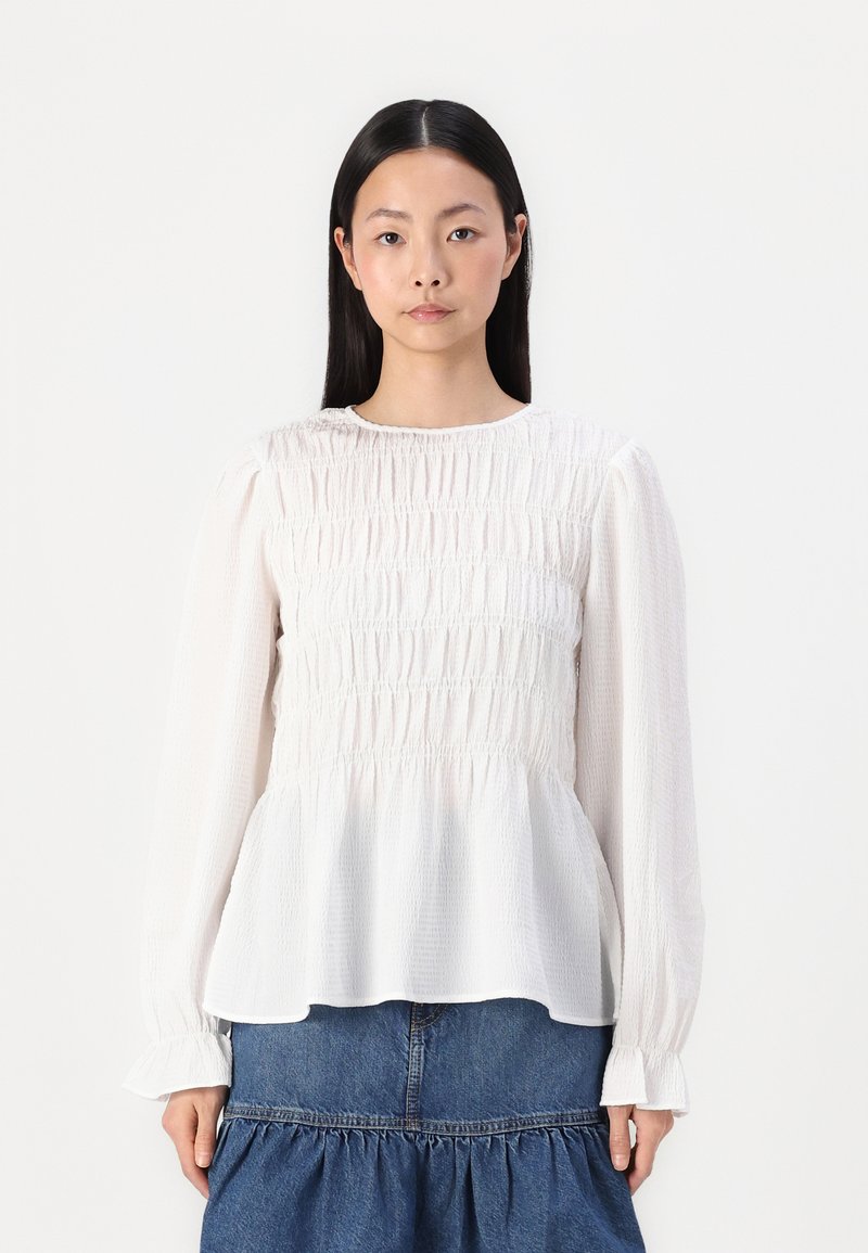 Hvit bluse med lange ermer, smocket midje og peplumnederkant. Har rund hals og volangmansjetter. Teksturert stoff gir detaljer.