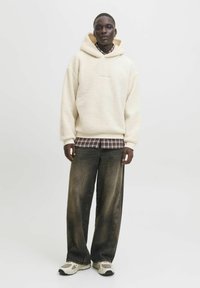 Crème-kleurige, fluffy fleece hoodie met een capuchon, gedragen over een geruit shirt. Gecombineerd met losvallende, vervaagde zwarte broek en sneakers.