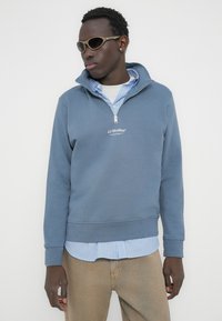 Άνδρας που φοράει μπεζ παντελόνι, μπλε φούτερ JJ Studios με φερμουάρ μέχρι το στήθος πάνω από ριγέ πουκάμισο με γιακά, γυαλιά ηλίου και ένα μικρό σκουλαρίκι, στέκεται μπροστά σε λευκό φόντο.