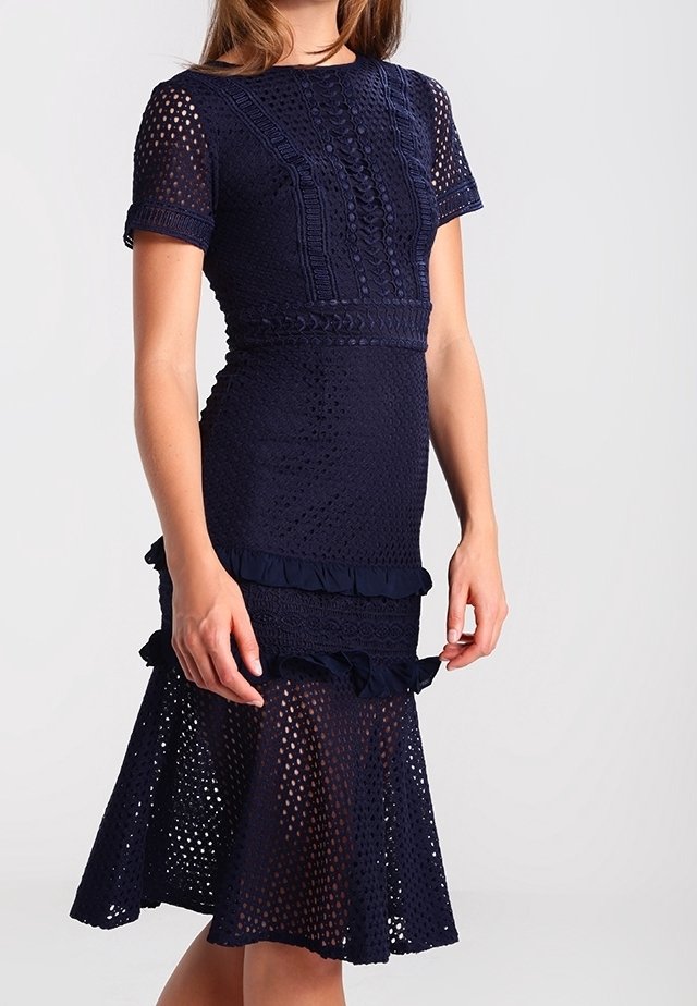 Robe en maille bleu marine à manches courtes, motifs texturés, détails à volants à la taille et à l’ourlet, portée par une femme debout sur un fond neutre.