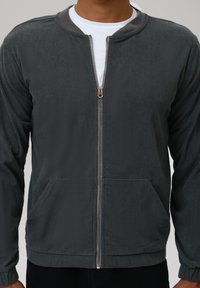 Jachetă zip-up din catifea gri cu guler rotund, având un fermoar de culoare aurie și buzunare laterale. Material texturat, croială casual.