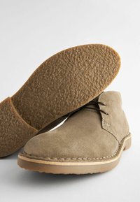 Botas desérticas de ante beige con detalles de costura, cordones oscuros y suelas de goma texturizada, mostradas con un zapato en posición vertical y la suela de otro zapato hacia adelante.
