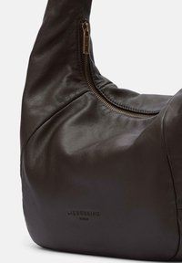 Braune Lederhandtasche mit glatter Oberfläche, ausgestattet mit einem Reißverschluss und einem geprägten Logo an der Seite. Abgerundetes Design und weiche Struktur.