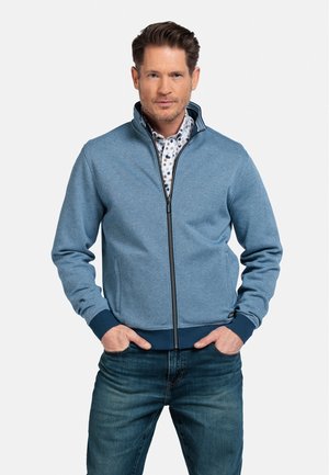 Lichtblauwe ritsjack met een textuurstof, marineblauwe accenten bij de mouwen en zoom, gedragen over een gestippeld shirt, gecombineerd met donkere jeans.