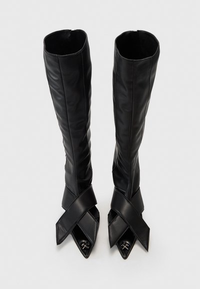 Botas negras hasta la rodilla hechas de cuero suave, con un diseño de correas entrecruzadas y puntas afiladas con acentos metálicos.