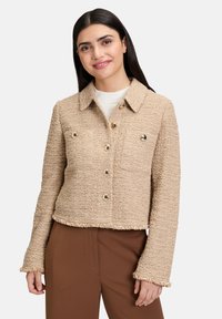 Giacca cropped beige in tessuto texture, con chiusura a bottoni, due tasche sul petto e orlo sfilacciato, abbinata a pantaloni marroni.
