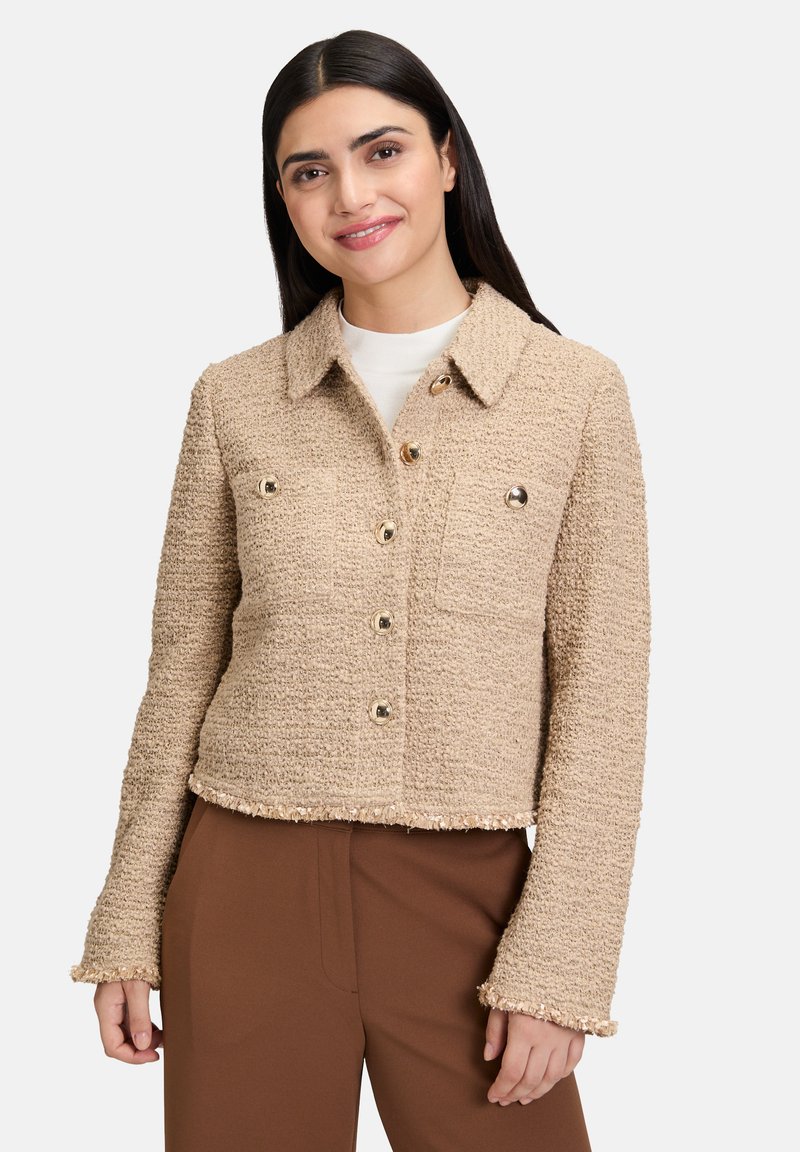 Giacca cropped beige in tessuto texture, con chiusura a bottoni, due tasche sul petto e orlo sfilacciato, abbinata a pantaloni marroni.