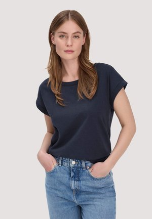 Junge Frau mit langen braunen Haaren, die ein marineblaues T-Shirt mit hochgekrempelten Ärmeln und hellblaue, hoch taillierte Jeans trägt, die Hände in den Taschen.