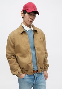 Világosbarna cipzáras dzseki rugalmas mandzsettával, kék pulóverre vetve, világoskék farmerrel és piros sapkával viselve. Minimalista dizájn, minták nélkül.