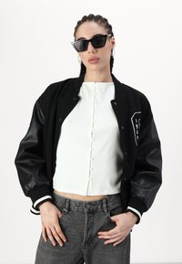 Veste varsity noire avec des manches en cuir, haut blanc à côtes avec des boutons à l'avant, jean en denim gris et lunettes de soleil noires angulaires.
